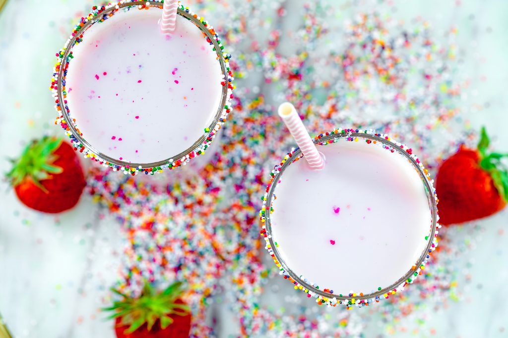 Overhead view of two glasses of pink strawberry milk on a marble tray with rainbow sprinkles and strawberries all around