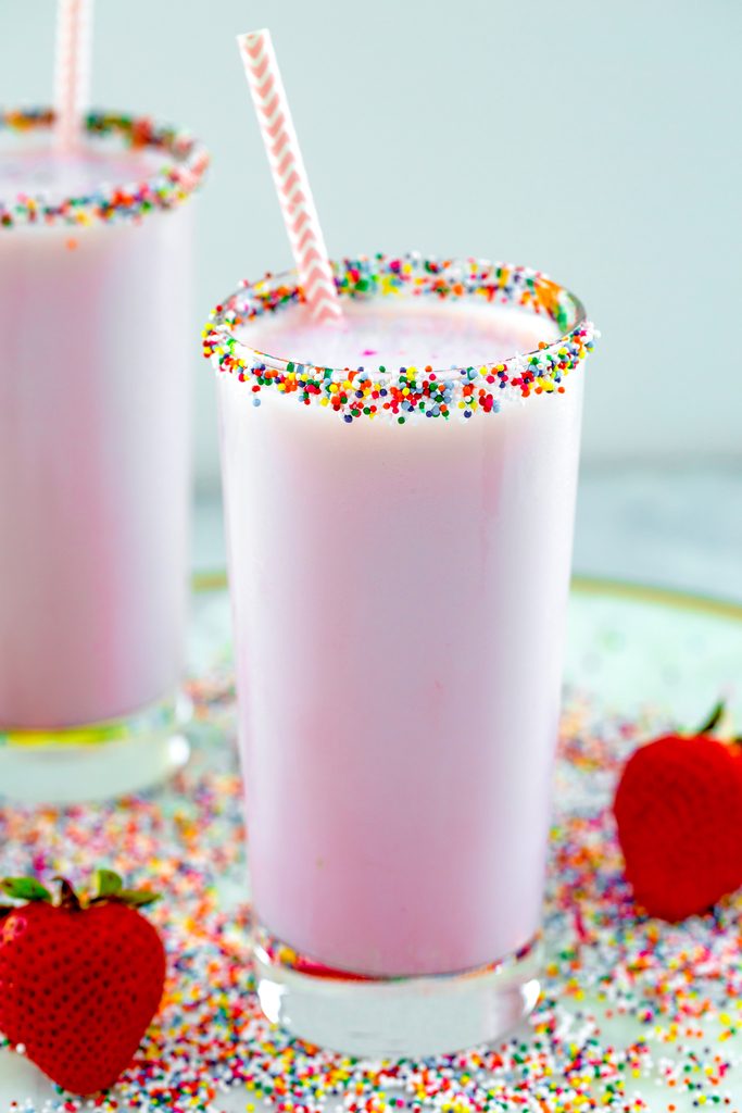 Head-on closeup view of a glass of pink strawberry milk with sprinkles on the rim with a second glass in the background and sprinkles and strawberries scattered around