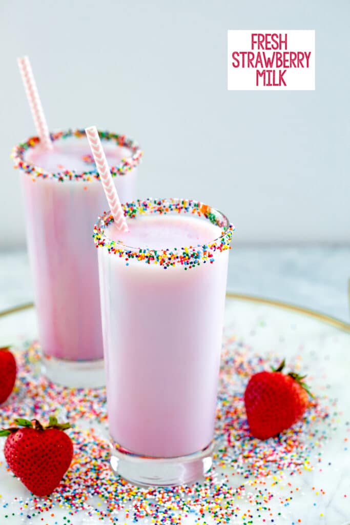 Head-on view of two glasses of pink strawberry milk with sprinkles on the rims on a marble tray with sprinkles and strawberries all around and recipe title at top
