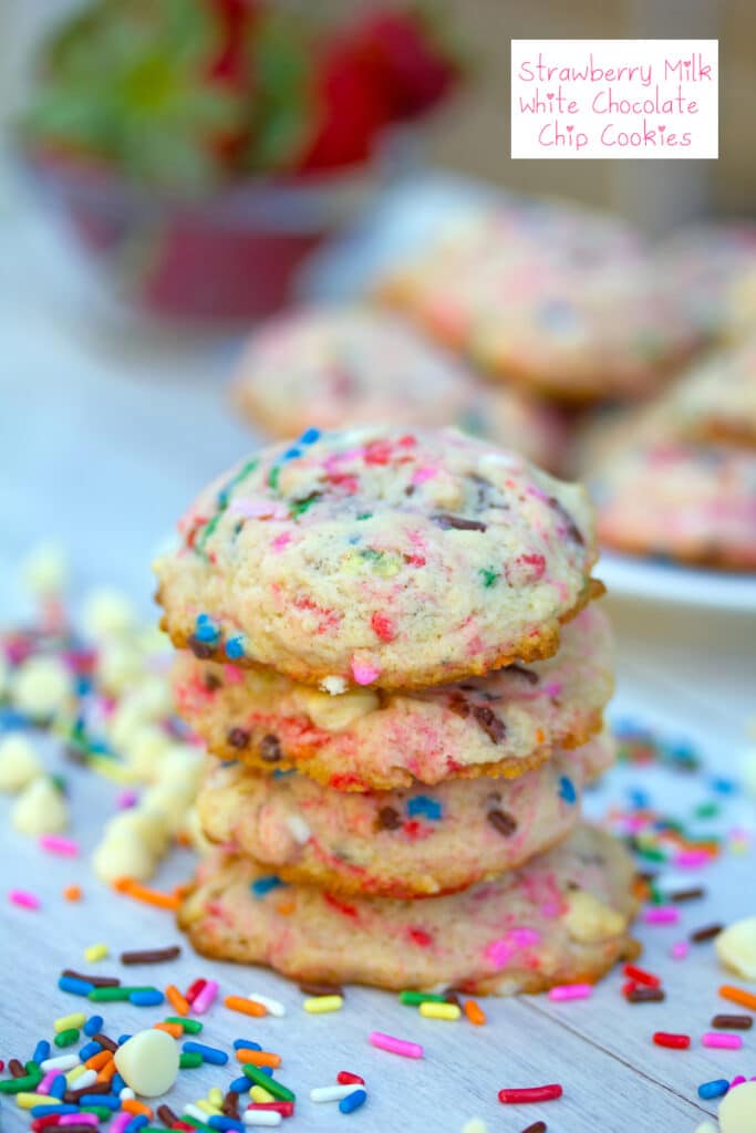 Head-on view of strawberry milk cookies surrounded by white chocolate chips and rainbow sprinkles with more cookies and strawberries in the background and recipe title at top