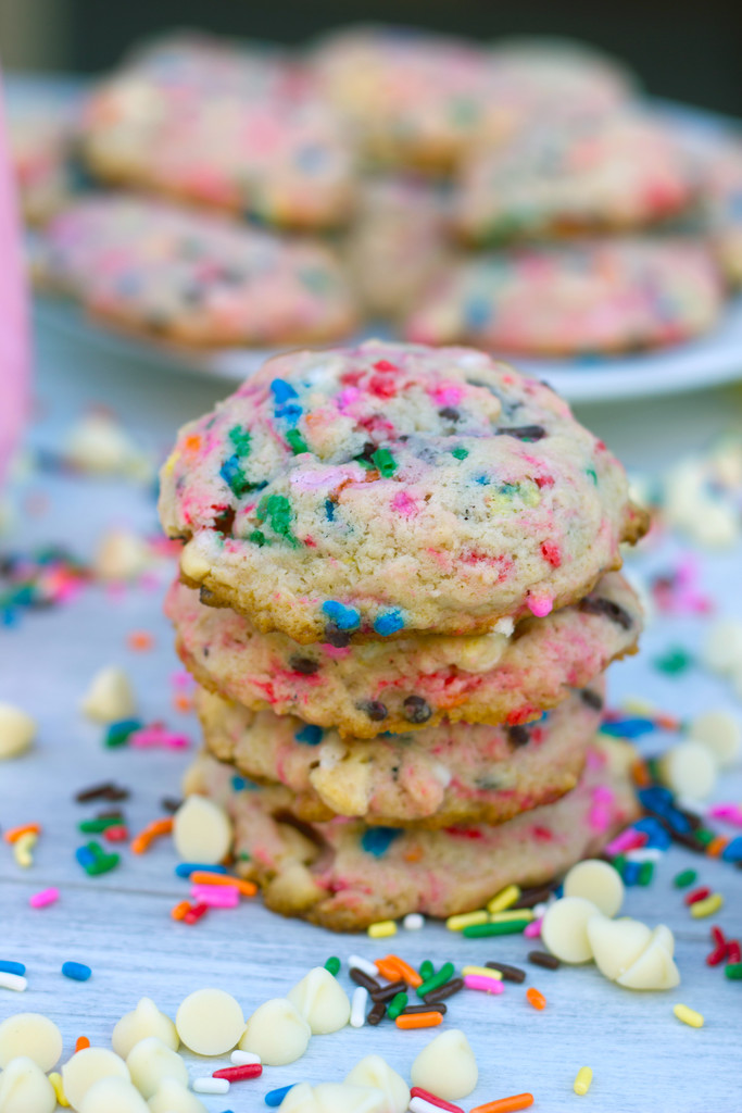 Close-up view of strawberry milk white chocolate chip cookies surrounded by white chocolate chips and rainbow sprinkles with platter of cookies in the background