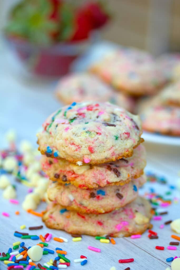 Head-on view of strawberry milk cookies surrounded by white chocolate chips and rainbow sprinkles with more cookies and strawberries in the background
