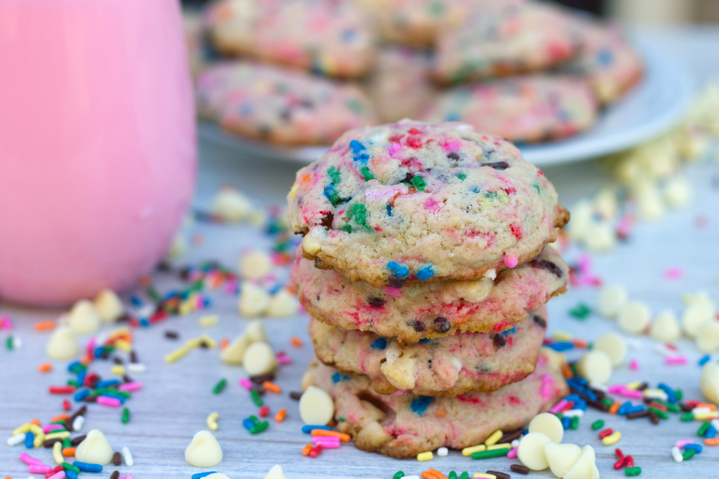 Landscape view of a stack of four strawberry milk cookies surrounded by white chocolate chips and rainbow sprinkles with platter of cookies and glass of strawberry milk in the background