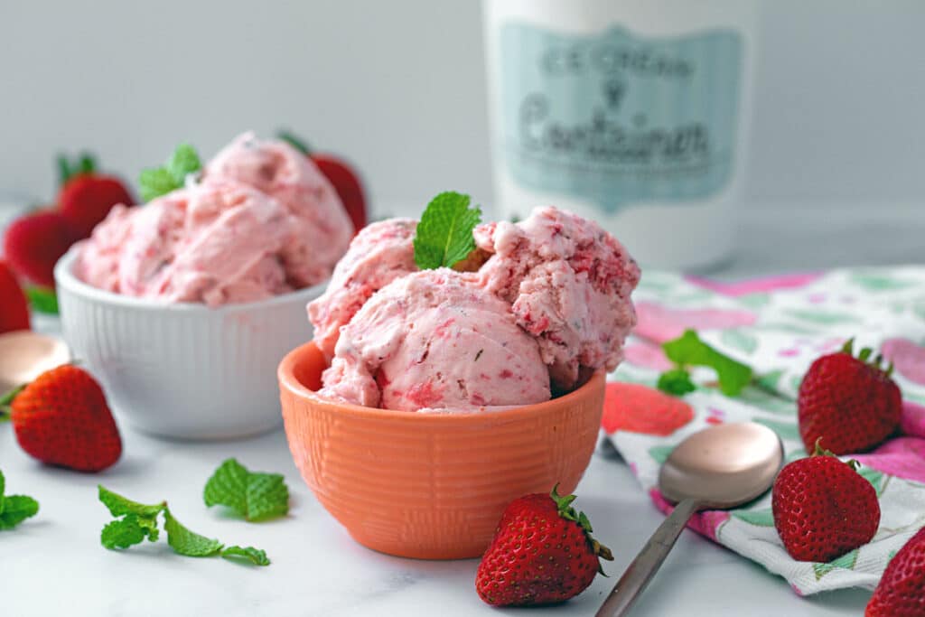 Landscape head-on view of two bowls of strawberry mint ice cream with quart container in background and whole strawberries and mint all around.