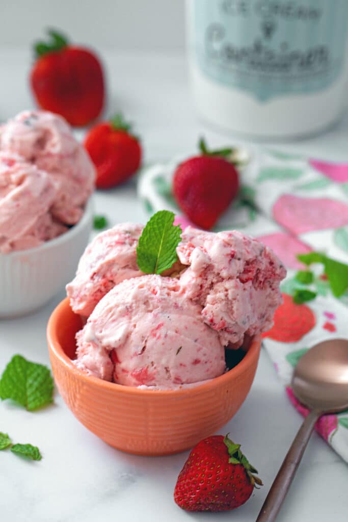 Head-on view of a bowl of strawberry mint ice cream with second bowl and spoon in background and strawberries and mint all around