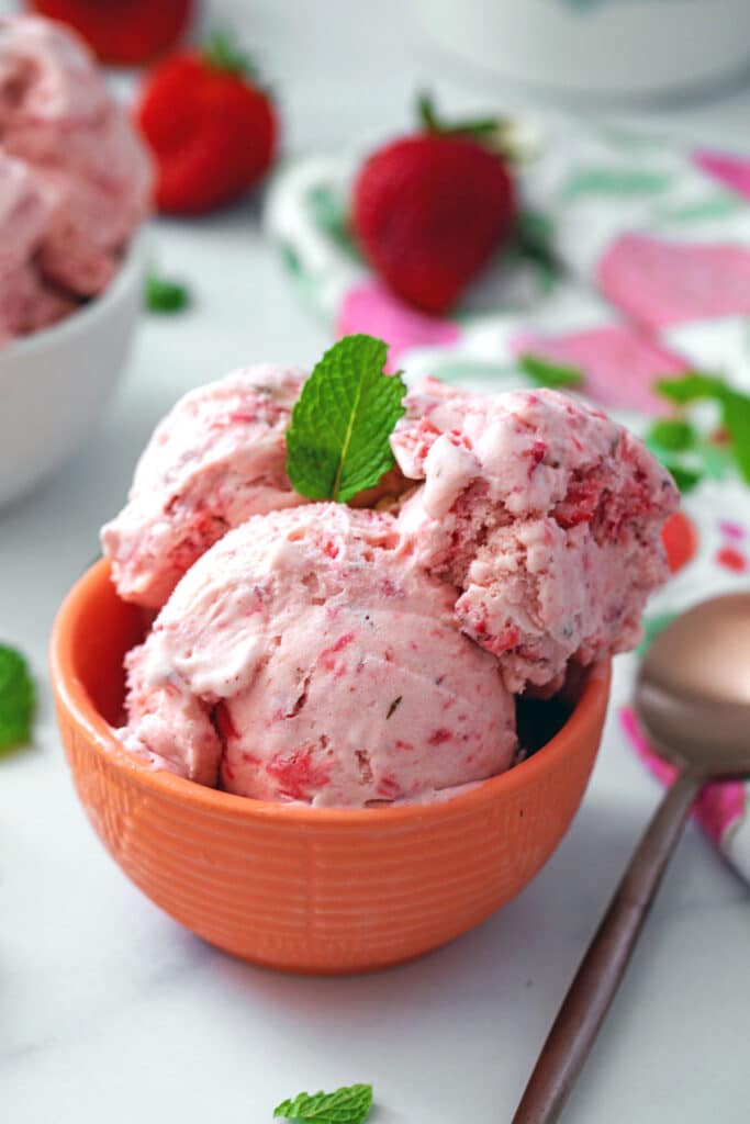 Pink strawberry mint ice cream in a bowl with mint leaf on top and strawberries all around
