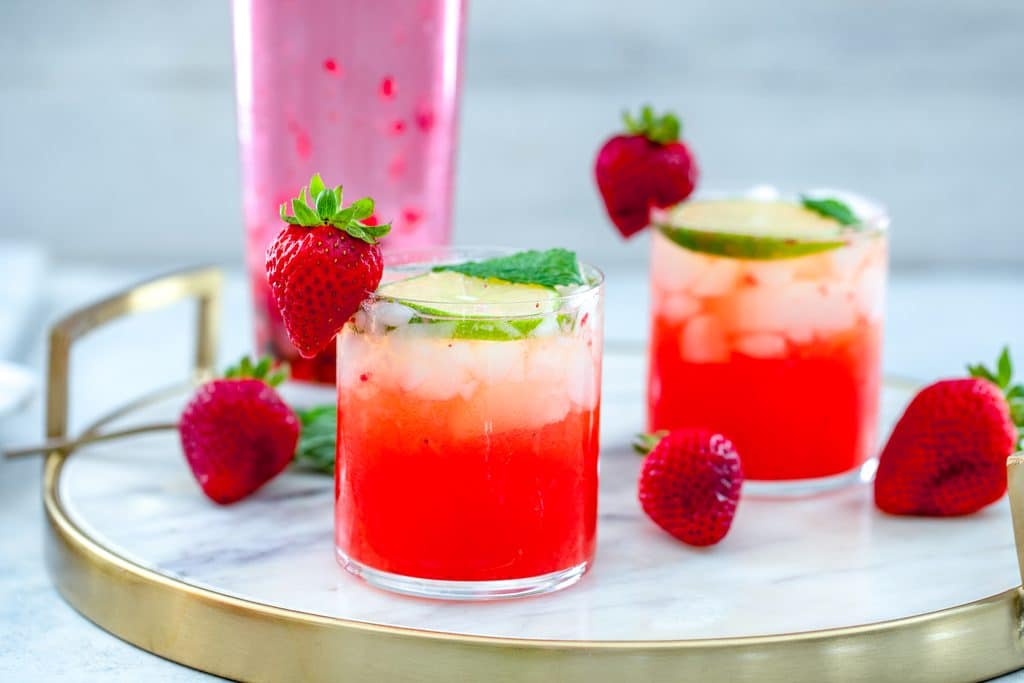 Landscape view of two strawberry mojitos on a marble tray with strawberries scattered around and a pink cocktail shaker in the background