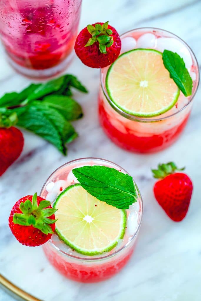 Bird's eye view of strawberry mojitos on a marble surface with strawberry, lime, and mint garnishes and mint and a pink cocktail shaker in the background