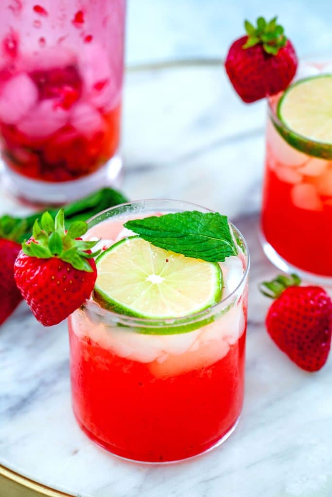 Head-on view of strawberry mojitos on a marble tray with lime, strawberry, and mint garnishes and a pink shaker in the background