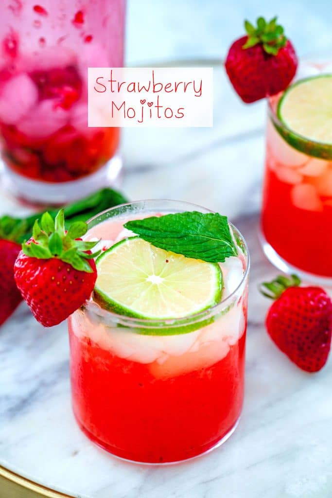 Head-on view of strawberry mojitos on a marble tray with lime, strawberry, and mint garnishes and a pink shaker in the background with "Strawberry Mojitos" text at top