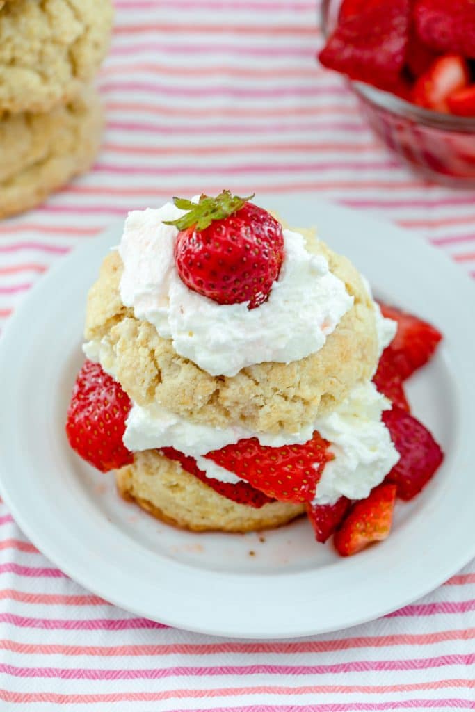 Overhead view of strawberry shortcake on a white plate with whipped cream and strawberries sandwiched between biscuits with whipped cream and a strawberry on top and more biscuits and bowl of strawberries in background