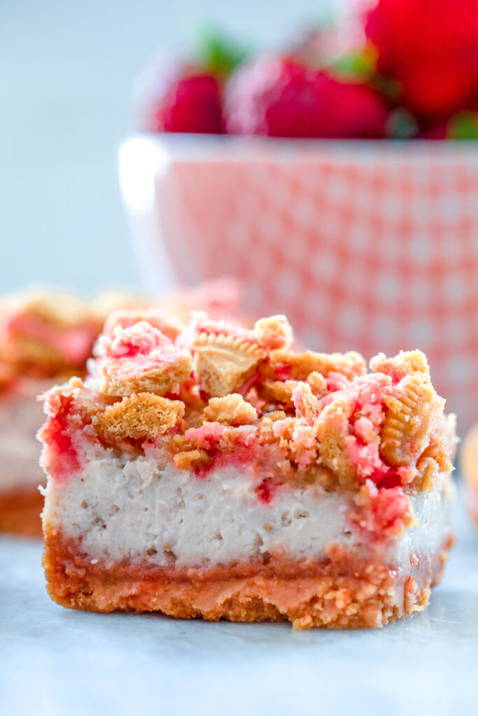 Up-close view of an strawberry shortcake cheesecake bar with red bowl of strawberries in the background