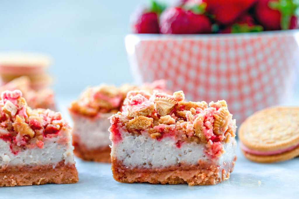 Landscape view of strawberry shortcake cheesecake bars with red bowl filled with strawberries and Oreo Cookie in the background