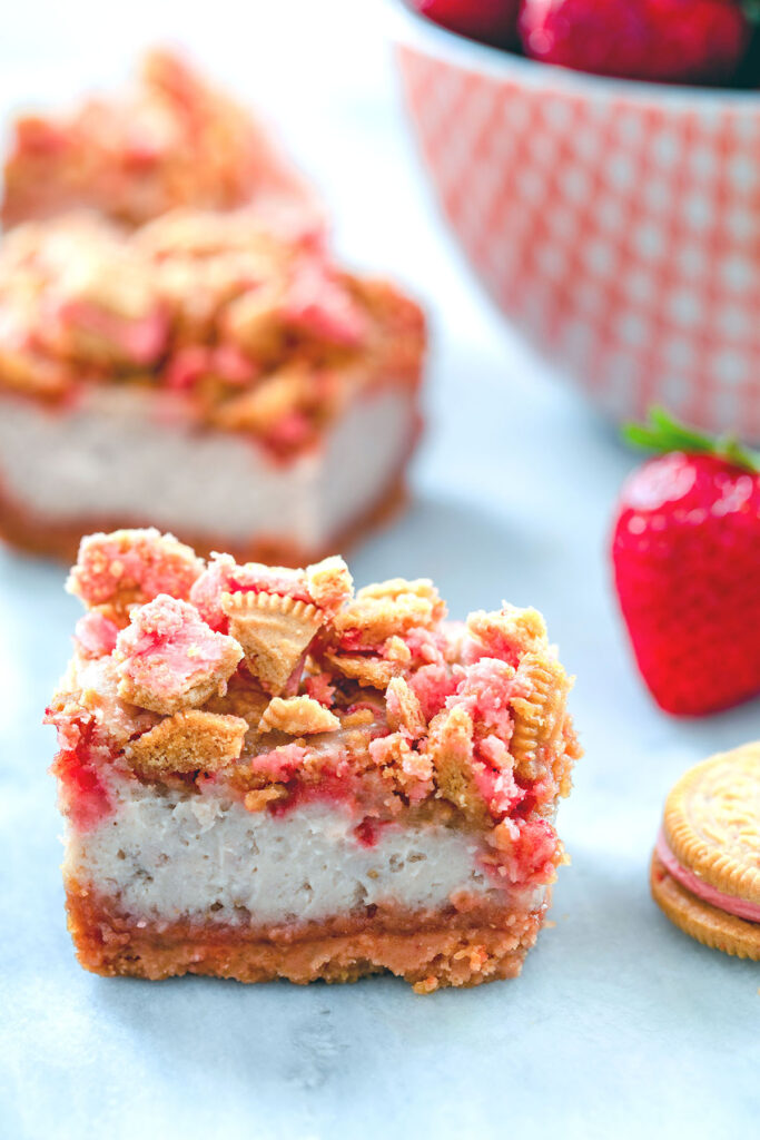 Head-on view of strawberry shortcake cheesecake bars with cookie crumbles and bowl of strawberries in the background