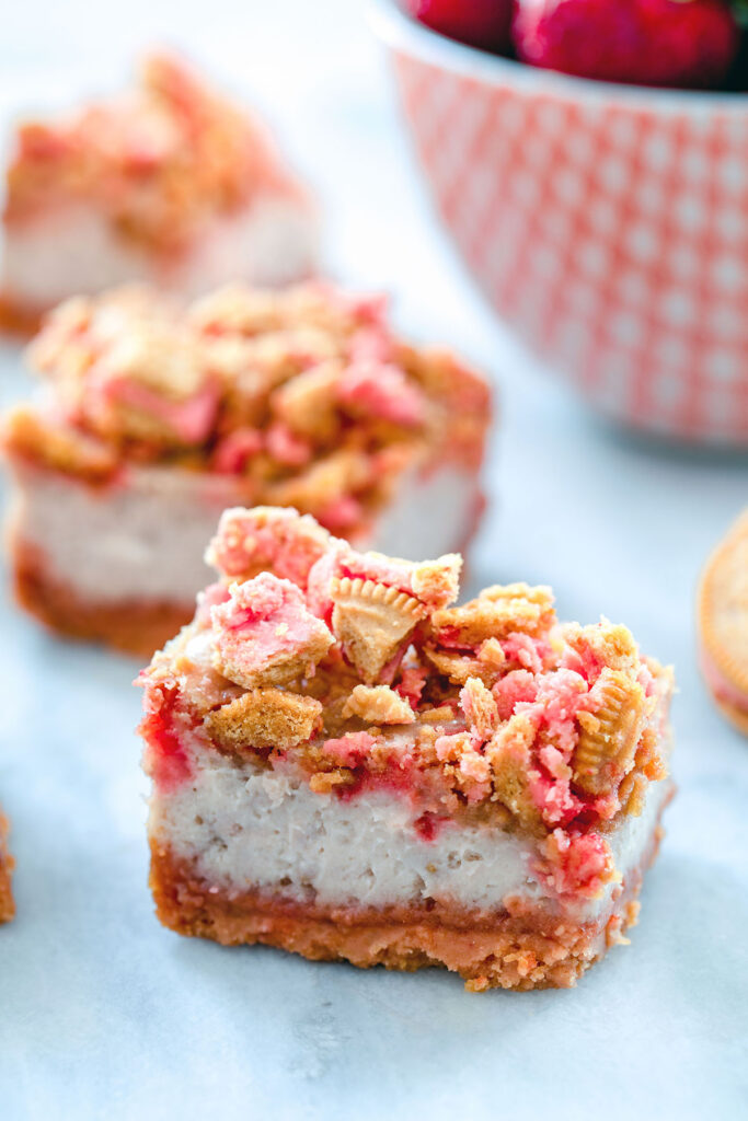 Up-close head-on view of strawberry shortcake cheesecake bar with more in the background and red bowl filled with strawberries