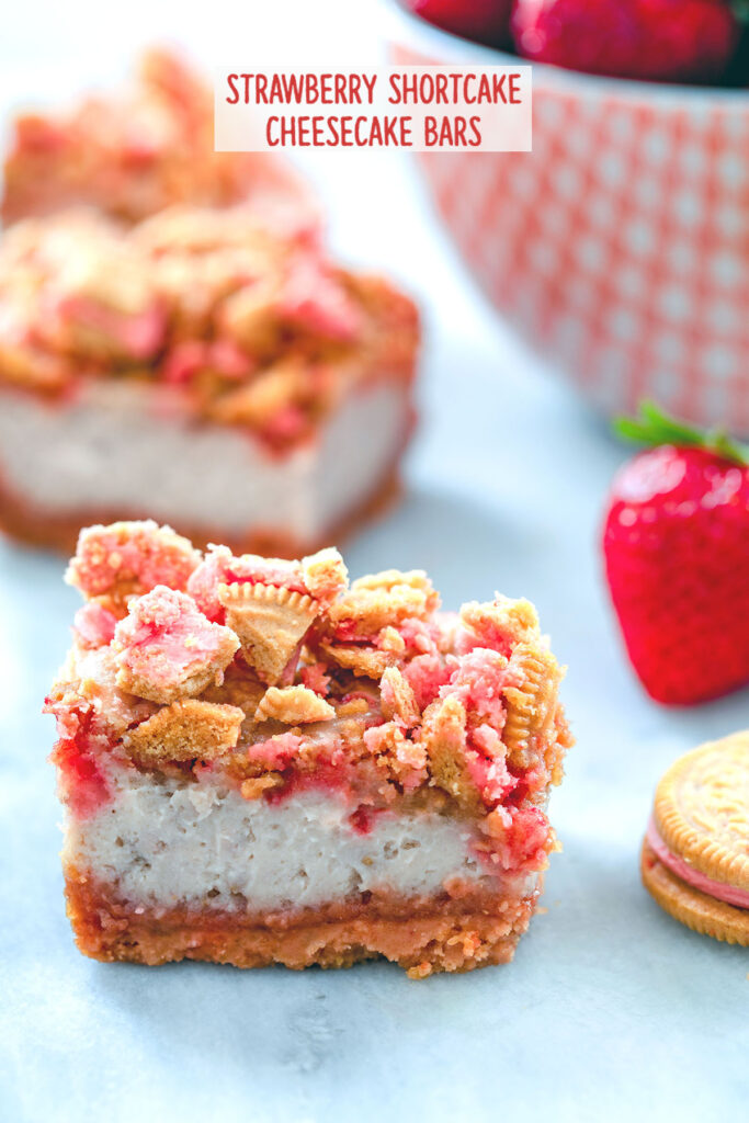 Head-on view of strawberry shortcake cheesecake bars with cookie crumbles and bowl of strawberries in the background with "Strawberry Shortcake Cheesecake Bars" text at top