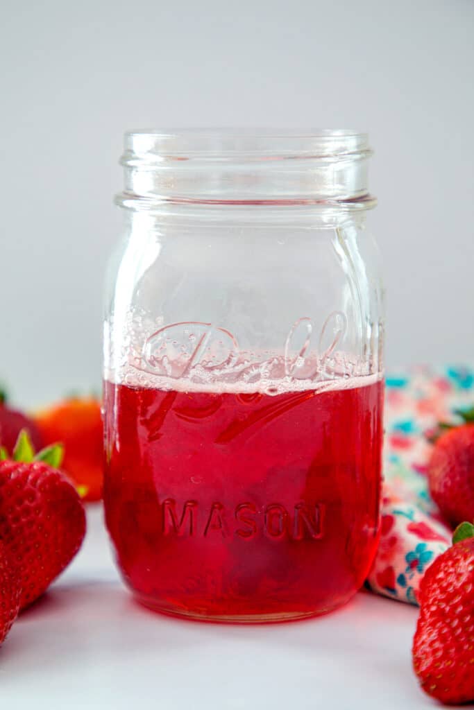 Head-on view of a mason jar of strawberry simple syrup with whole strawberries all around.