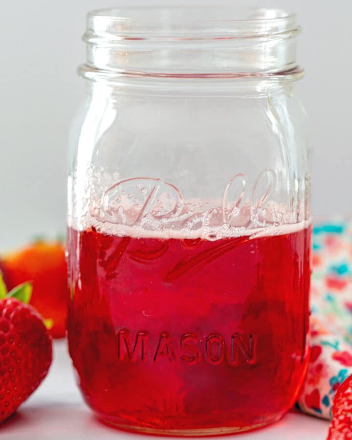 Head-on closeup view of a mason jar of strawberry simple syrup with whole strawberries all around