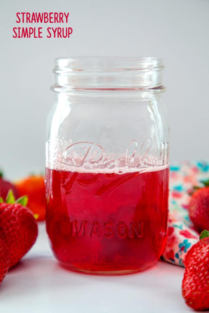 Head-on view of a jar of bright red strawberry simple syrup with strawberries all around.