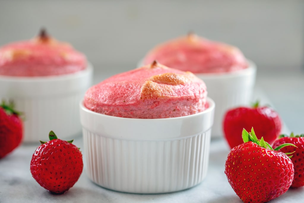Head-on landscape view of a pink strawberry souffle in a white ramekin surrounded by whole strawberries with more souffles in the background
