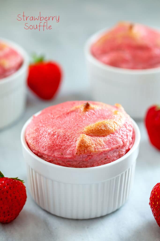 Overhead view of a pink strawberry souffle in a white ramekin with more souffles and whole strawberries in the background with recipe title at top
