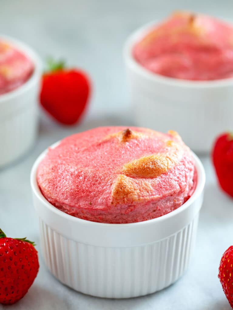 Head-on view of a strawberry souffle in a white ramekin with more souffles in background and strawberries all around.