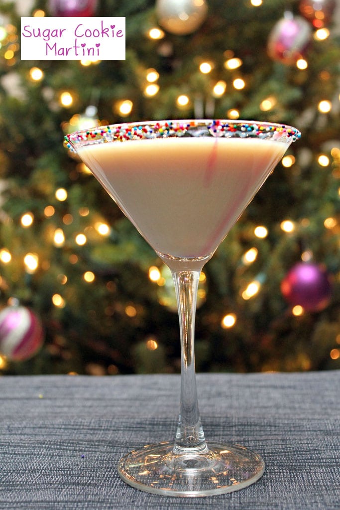 Head-on view of a sugar cookie martini with sprinkles rim in front of a lit Christmas tree with recipe title at top