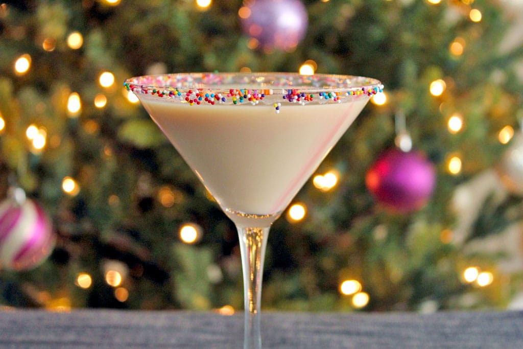 Landscape head-on view of sugar cookie martini with sprinkles rim in front of a lit Christmas tree