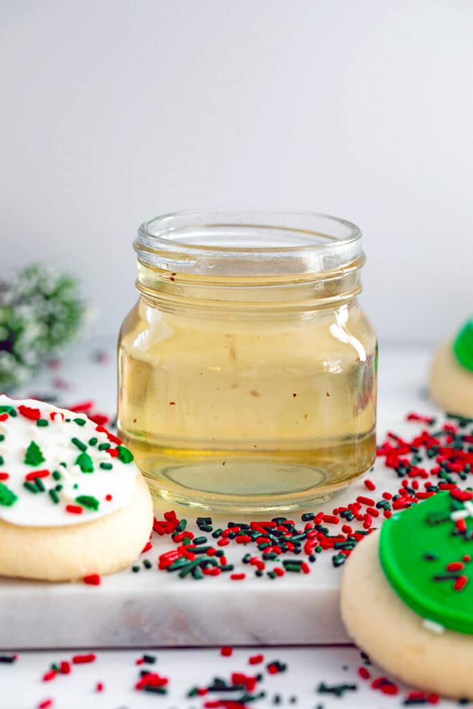 Head-on view of small jar of sugar cookie syrup with red and green sprinkles and Christmas cookies all around.