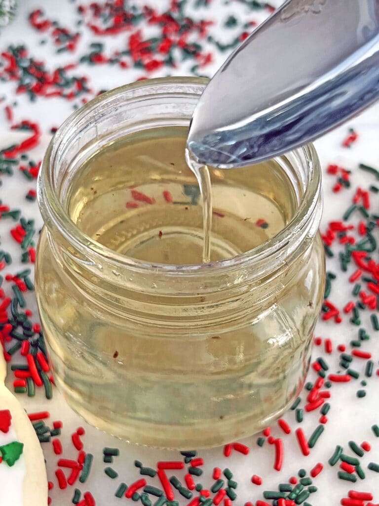 Overhead view of a small jar of sugar cookie syrup with some drizzling off a spoon with red and green sprinkles all around.