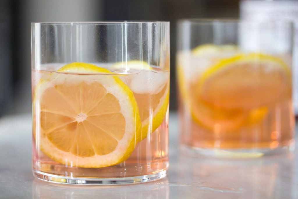 A landscape photo featuring two rocks glasses of pink rosé spritzer with lemon rounds on a white marble surface