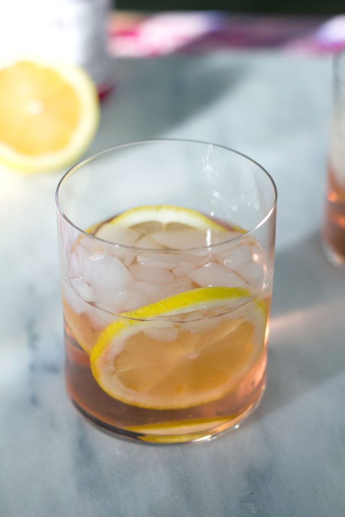 A from-above view of pink summer rosé spritzer in a rocks glass with lemon rounds and plenty of crushed ice and a lemon half in the background