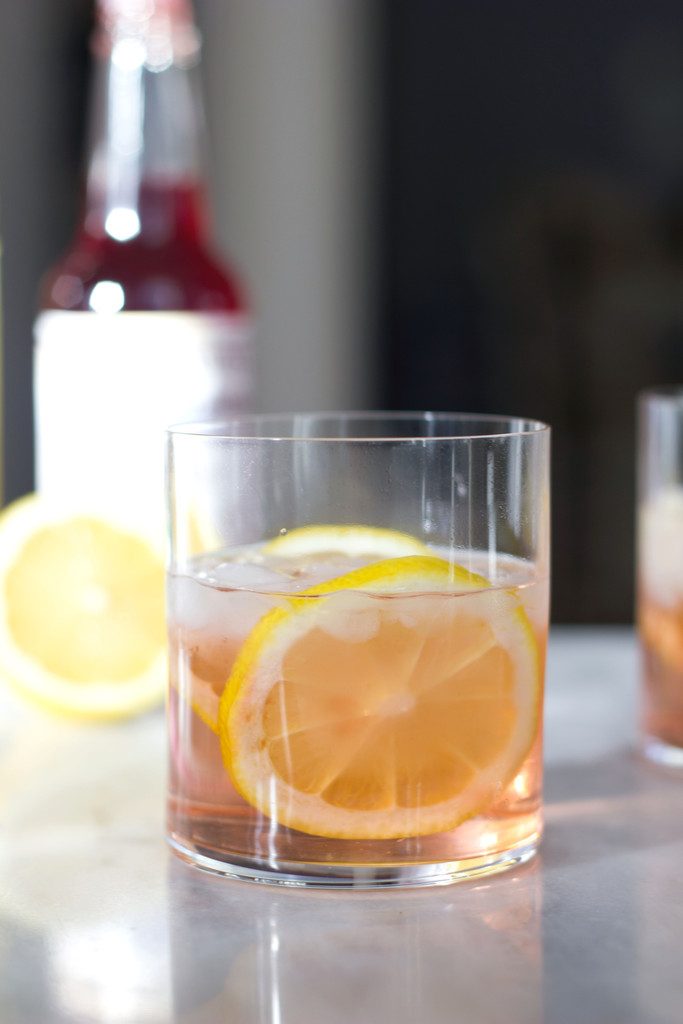 A head-on view of a pink rosé spritzer in a rocks glass with lemon rounds and a lemon half and bottle of bitters in the background