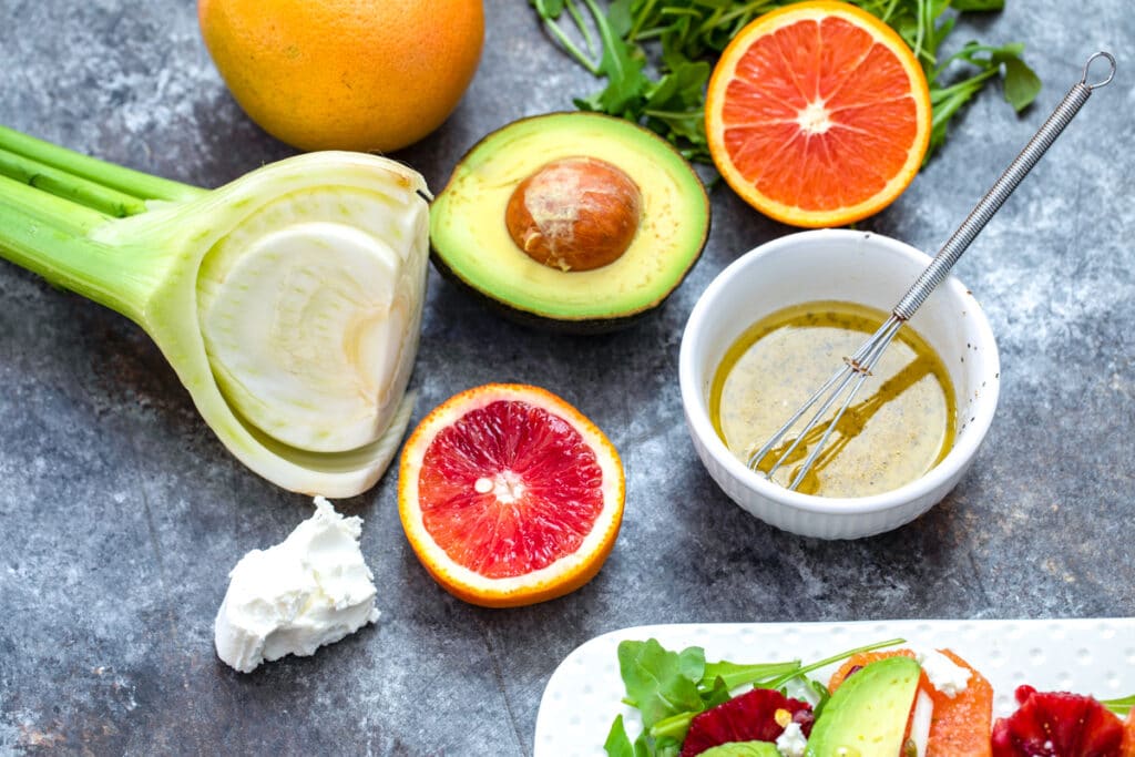 Overhead view of ingredients in salad, including fennel bulb, half an avocado, half a blood orange and grapefruit, hunk of goat cheese, arugula, and dressing in a white bowl with small whisk
