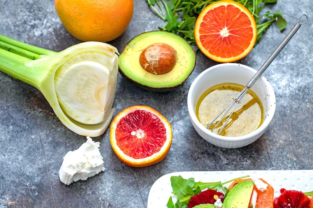 Overhead view of ingredients in salad, including fennel bulb, half an avocado, half a blood orange and grapefruit, hunk of goat cheese, arugula, and dressing in a white bowl with small whisk