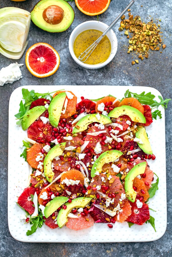 Overhead view of winter citrus salad on a white platter with pistachios, blood orange half, grapefruit half, goat cheese, fennel bulb, half an avocado, and small white bowl of dressing