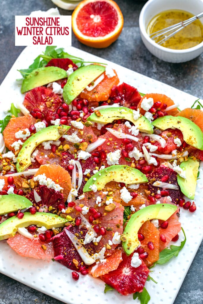 Overhead close-up view of winter citrus salad with blood oranges, cara cara oranges, grapefruit, fennel, avocado, goat cheese, and more with ingredients in the background and recipe title at top