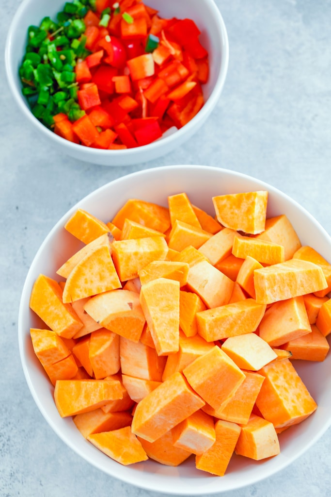 Overhead view of chopped and cubed sweet potatoes with bowl of chopped red peppers and jalapeños in the background