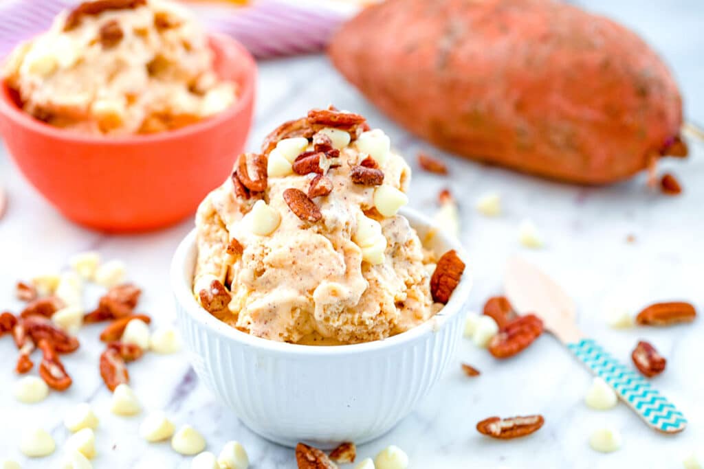Landscape head-on view of a bowl of sweet potato ice cream with white chocolate chips and toasted pecan with second bowl of ice cream, small wooden spoon, and whole sweet potato in the background