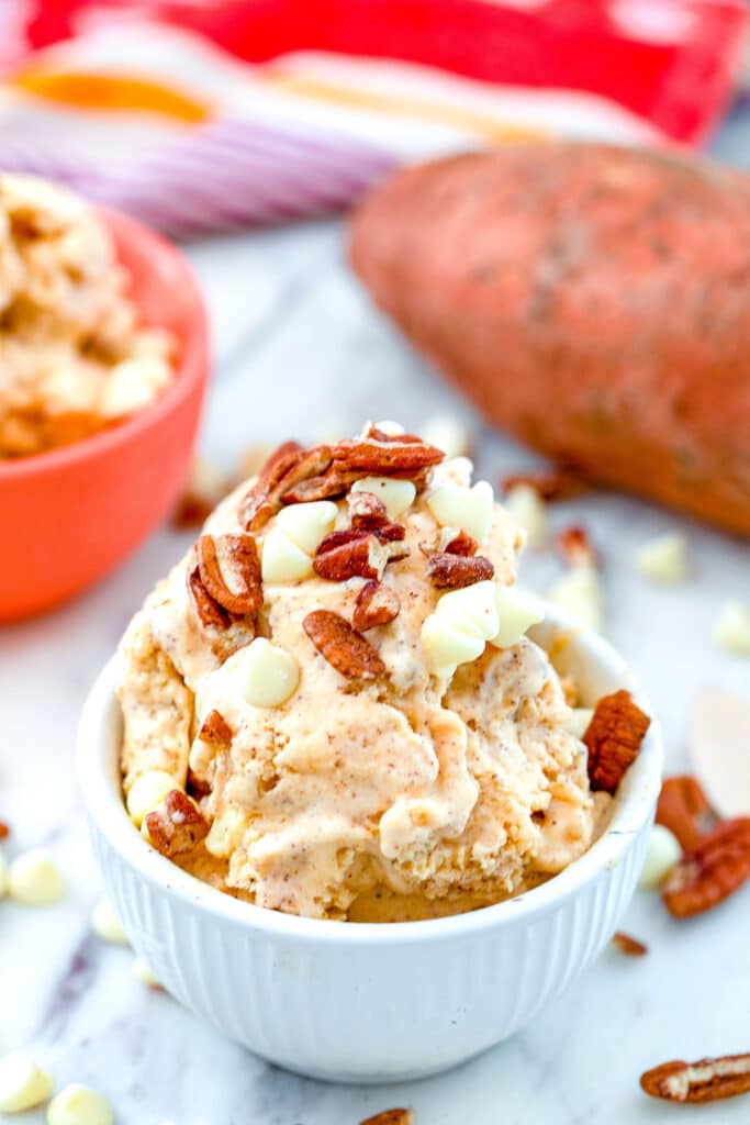 Head-on view of a white bowl of sweet potato ice cream topped with white chocolate chips and toasted pecans with second bowl of ice cream and sweet potato in background