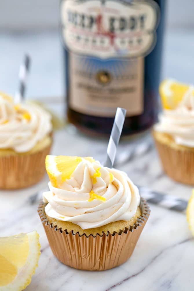 Head-on photo of sweet tea vodka cupcake with a grey and white straw, lemon wedge, and lemon zest garnish, other cupcakes and bottle of sweet tea vodka in background