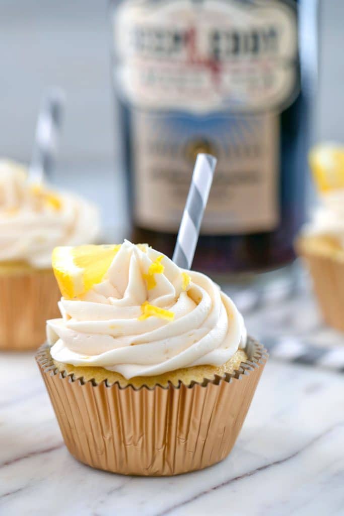 Up-close view of sweet tea vodka cupcake topped with a grey and white striped straw, lemon wedge and lemon zest with a bottle of sweet tea vodka in the background