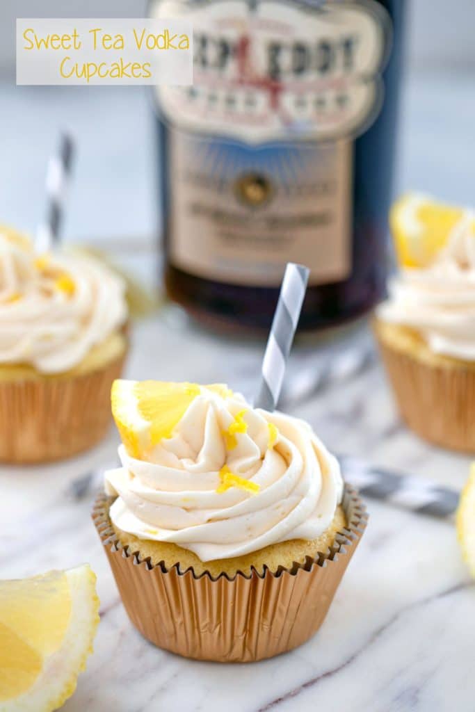 Head-on photo of sweet tea vodka cupcake with a grey and white straw, lemon wedge, and lemon zest garnish, other cupcakes and bottle of sweet tea vodka in background, and "Sweet Tea Vodka Cupcakes" text at top