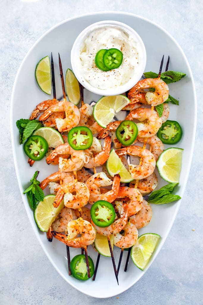 Bird's eye view from distance showing white platter with grilled tequila shrimp skewers with jalapeño slices, lime wedges, and yogurt dip in small bowl.