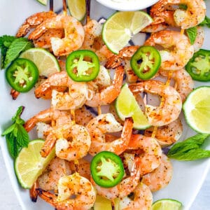 Bird's eye closeup view of white platter with skewers of tequila shrimp with sliced jalapeños, lime wedges, and yogurt dip