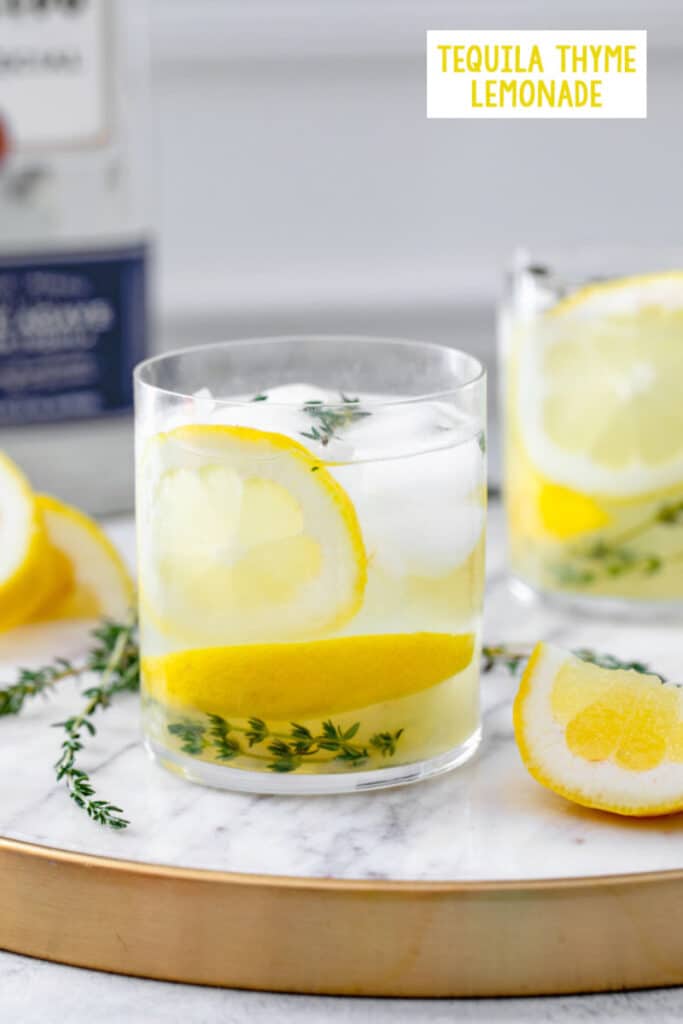 Head-on close-up view of a glass of tequila thyme lemonade with lemon rounds and fresh thyme with more lemons and thyme in background and recipe title at top
