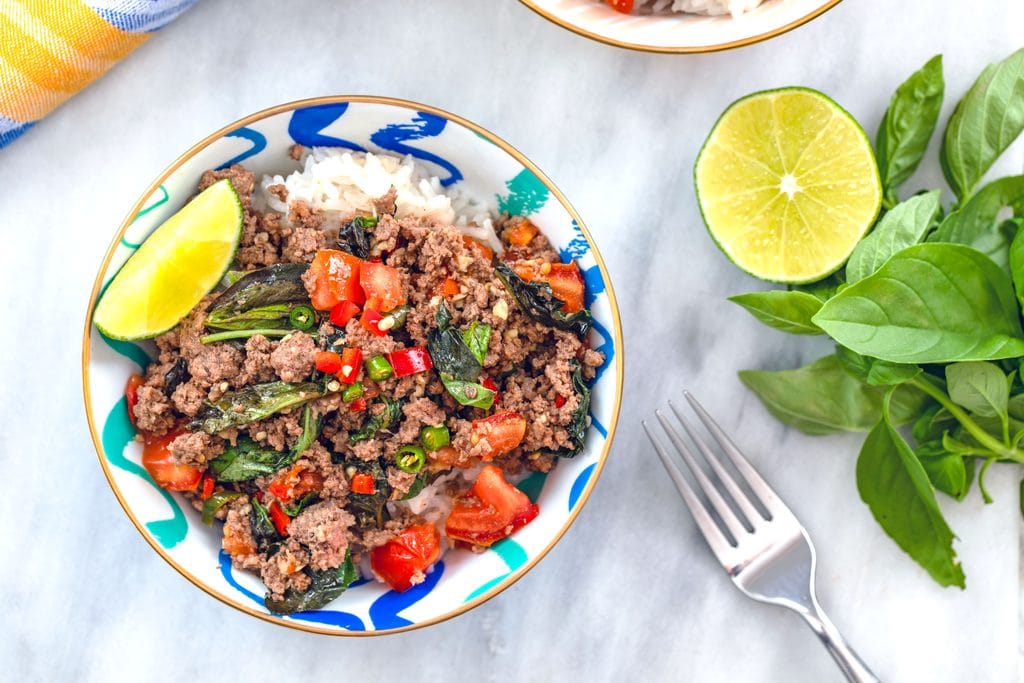 Landscape view of bowl of Thai beef basil served over coconut rice on a marble surface with fork, basil, and half a lime next to bowl