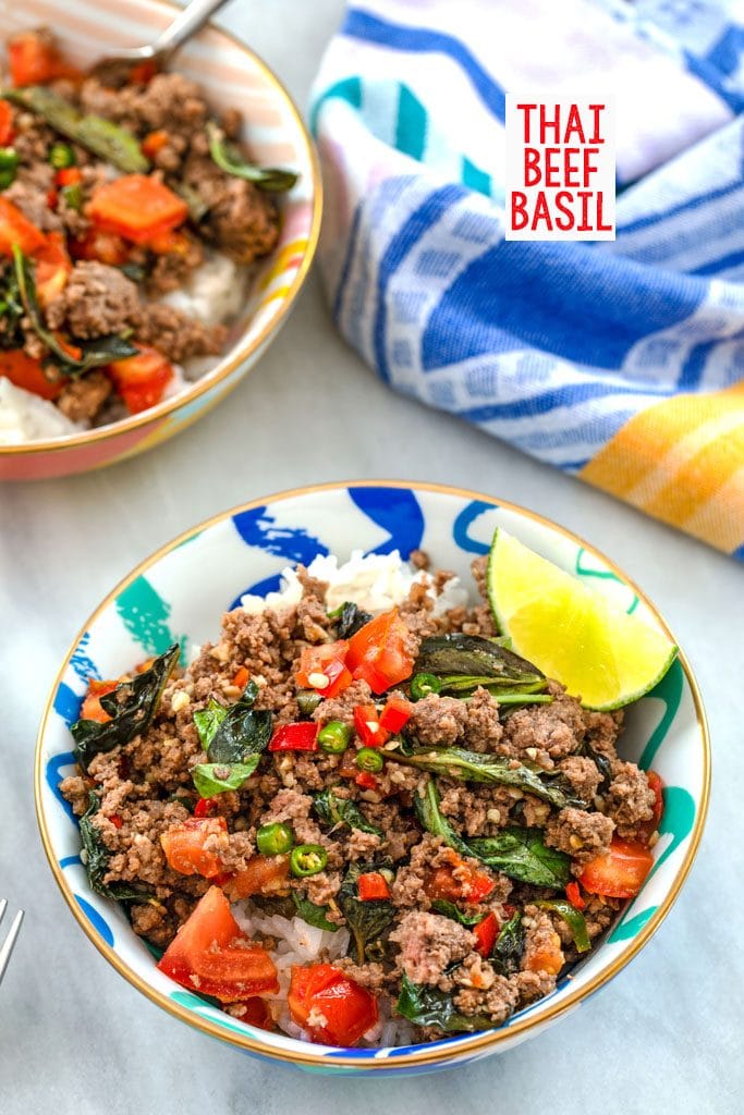 Overhead view of bowl of Thai beef basil served over coconut rice with a lime wedge, bowl in the background, and colorful dish towel with "Thai Beef Basil" text at top