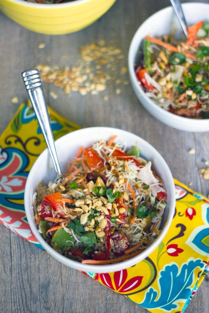 Overhead view of two white bowls of Thai quinoa salad with peanut dressing on a colorful napkin with bowl of dressing indn the background and peanuts scattered arou