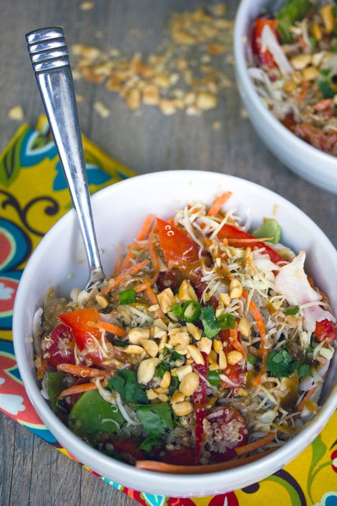 Overhead view of a white bowl of Thai quinoa salad with a fork on a colorful napkin with second bowl in the background and chopped peanuts all around